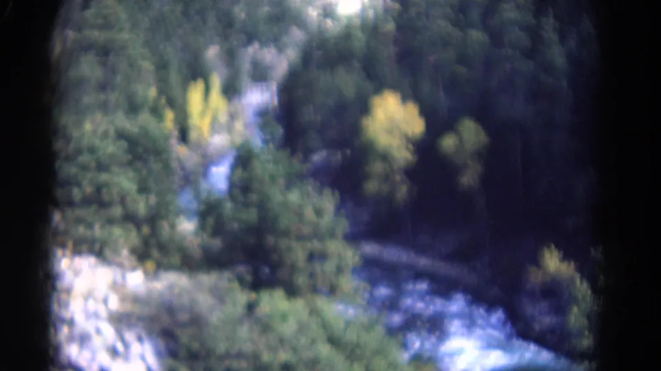 1967 Colorado landscape frame with a pale river channel, dense trees, yellow autumn foliage, and rocky valley terrain.