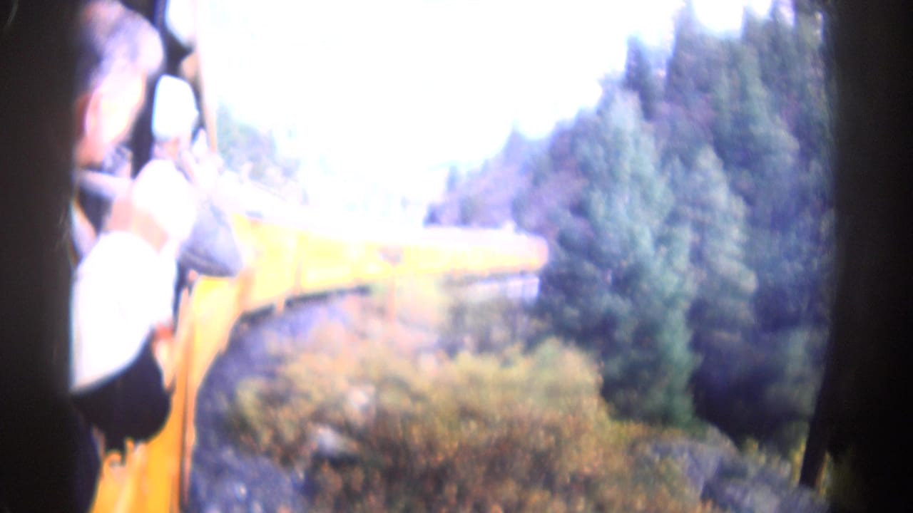 1967 Colorado home-movie frame showing tourists at open train windows beside yellow rail cars in a wooded mountain setting.