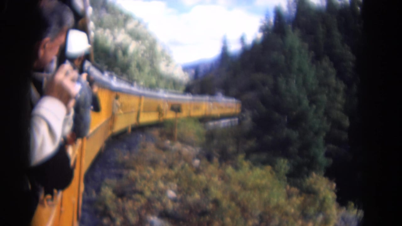 1967 Colorado 8mm frame with a passenger photographing from an open train window as yellow rail cars bend through trees.