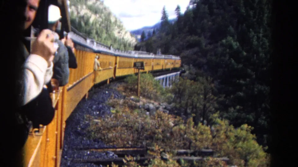 1967 Colorado 8mm frame showing yellow passenger cars curving past trees while a tourist photographs from the foreground window.