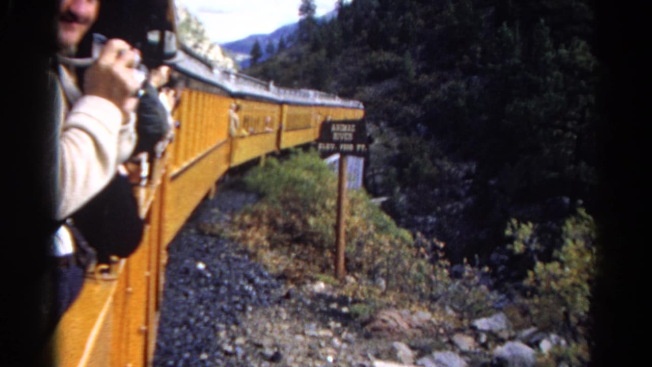 1967 Colorado 8mm frame showing a passenger photographing from a yellow train near forested mountains and a trackside sign.
