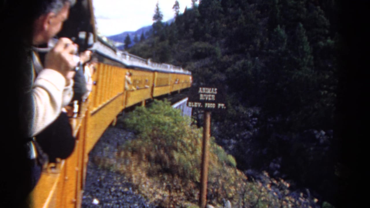 1967 Colorado train-view frame with a yellow passenger car, camera-holding rider, wooded slope, and Animas River elevation sign.