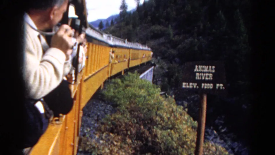 1967 Colorado 8mm frame showing the Animas River elevation sign beside a moving yellow train and mountain forest.