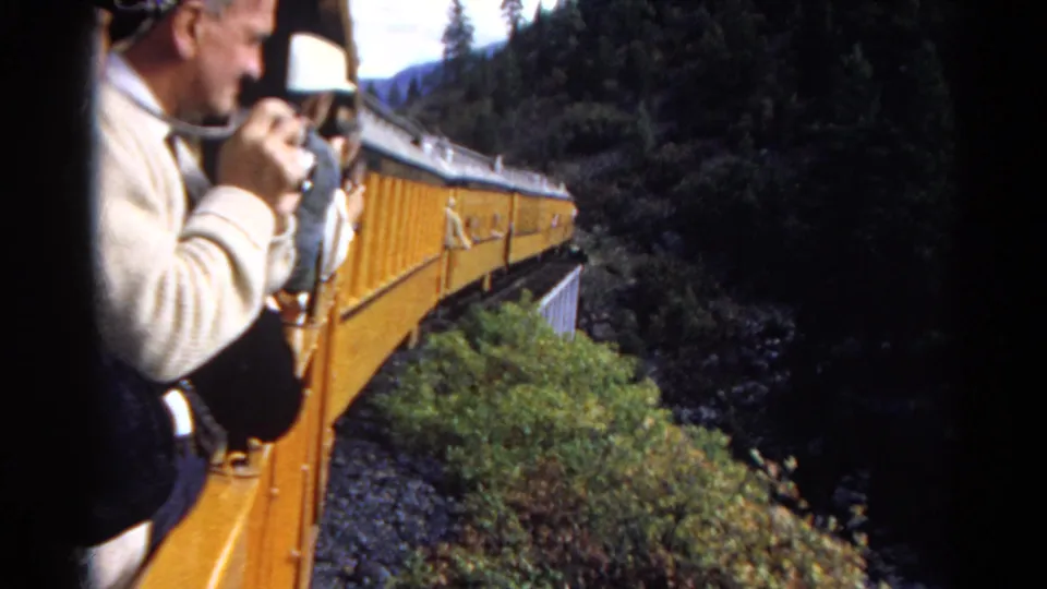 1967 Colorado travel frame showing a passenger leaning from a yellow train near a trestle and wooded mountain slope.