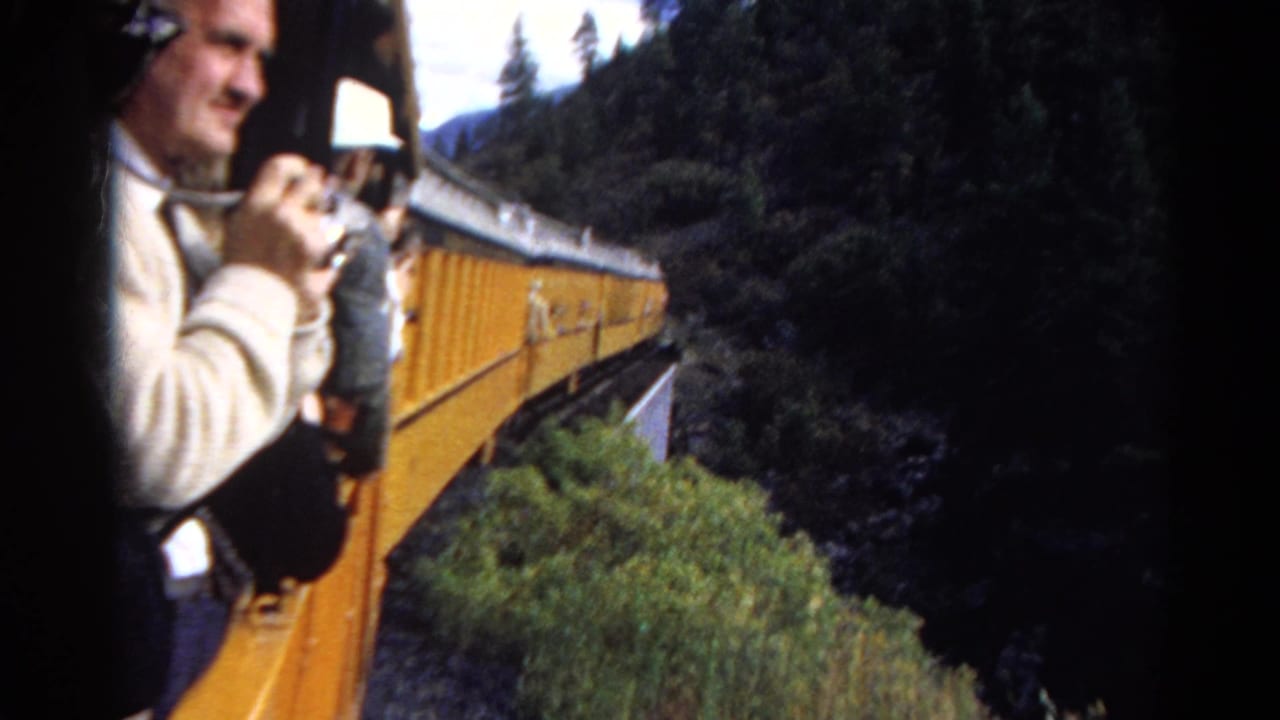 In 1967 Colorado home-movie footage, an adult man with a camera leans from a yellow passenger train as forested slopes pass nearby.