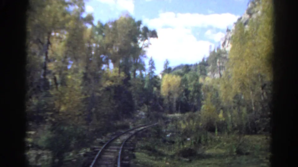 A passenger-view shot shows rails curving through Colorado forest and brush with rocky slopes and bright sky beyond.