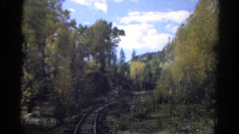 The train route continues through wooded Colorado mountain terrain, with rails curving into trees beneath a partly cloudy sky.