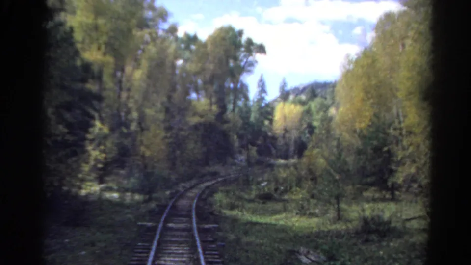 Rear view from a moving train in Colorado, showing tracks bending through a wooded valley with distant hills.