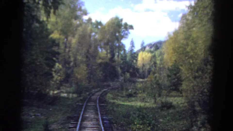1967 8mm rear train view with rails, ties, and wooded Colorado valley scenery softened by motion.