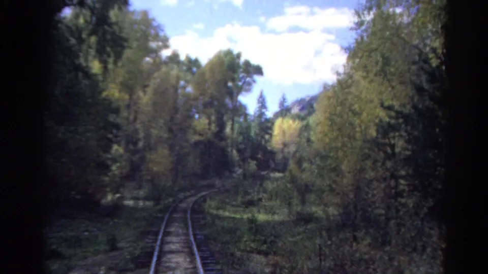 Rear-facing Colorado rail footage showing tracks disappearing into a wooded curve beneath a bright sky.