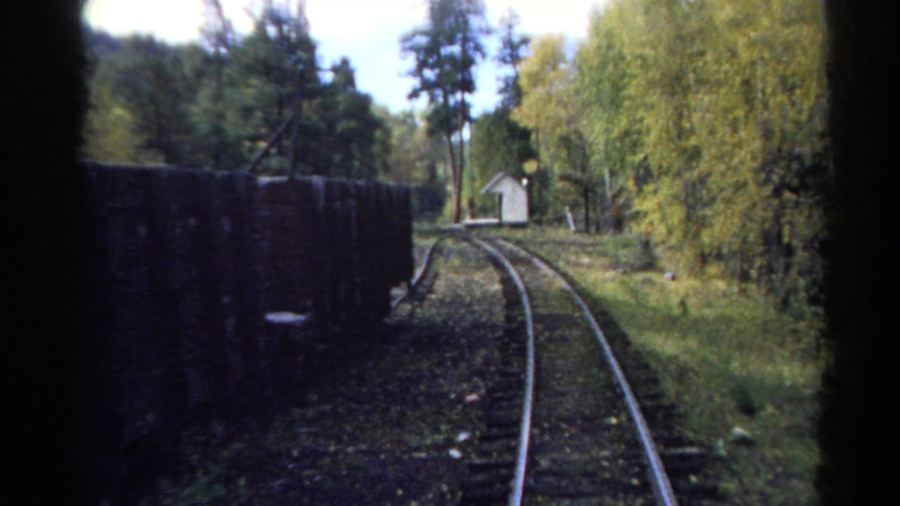 1967 Colorado 8mm view from a moving train, with curving rails, dark freight cars, green trees, and a small white building ahead.
