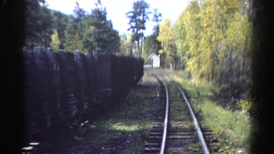 Colorado home-movie frame from a train, with rails bending past dark freight cars, dense trees, and a white structure beside the route.