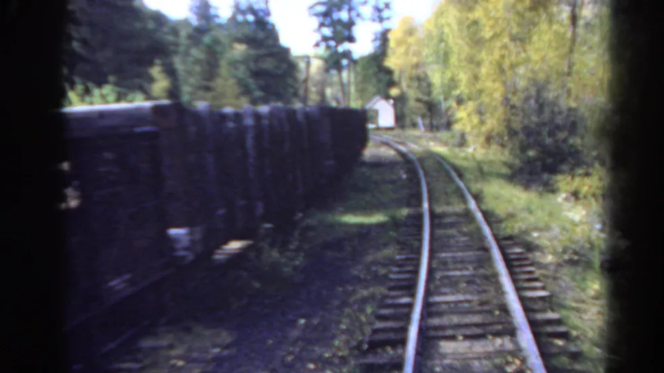 End frame of the 1967 Colorado train journey, showing a dark freight car close to the camera, curving rails, trees, and a small white building ahead.