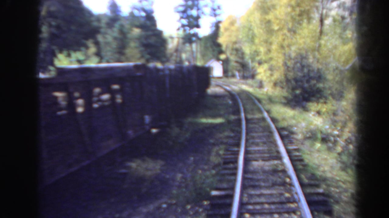 1967 Colorado 8mm frame from a moving train, showing curving rails, dark freight wagons, trees, grass, and bright sky.