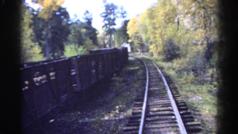 1967 Colorado home-movie frame showing a train-side view of freight wagons, rails, trees, grass, and soft 8mm motion blur.