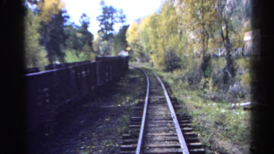 Colorado 1967 8mm still with curving rails, dark freight wagons, wooded slopes, yellow-green foliage, and a bright overexposed sky.
