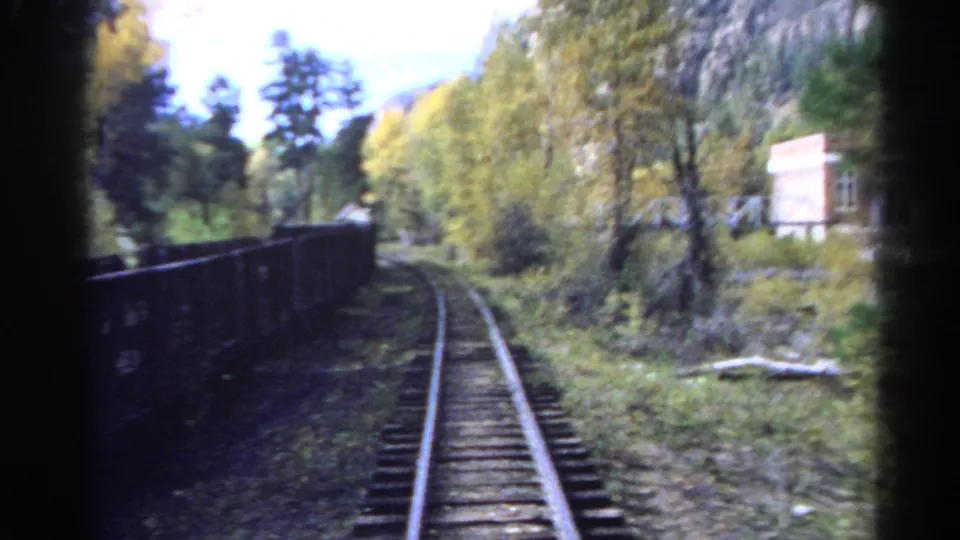 1967 Colorado 8mm frame looking down railroad tracks through autumn trees, with rocky mountains and a river corridor nearby.