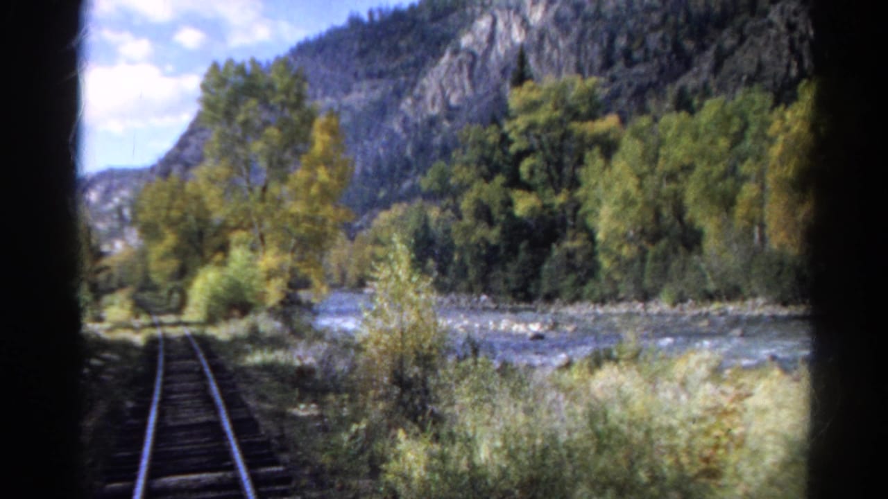 Railroad track runs beside a river with trees and rocky mountains beyond.