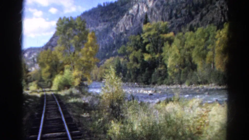 1967 Colorado rail-view frame showing tracks beside a river, dense trees, and steep rocky mountain slopes under a bright sky.
