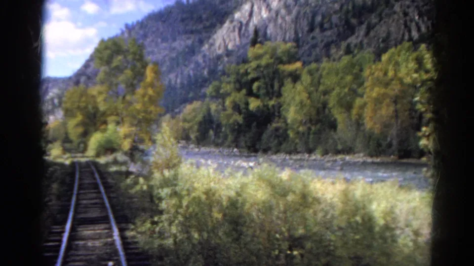 1967 Colorado 8mm frame of tracks running alongside a river, with brush, autumn trees, and rocky mountain terrain ahead.