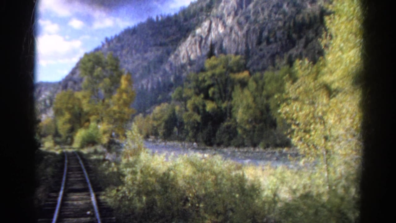 1967 Colorado 8mm frame from a moving train showing tracks, riverside trees, rocky mountains, and dark film-gate edges.