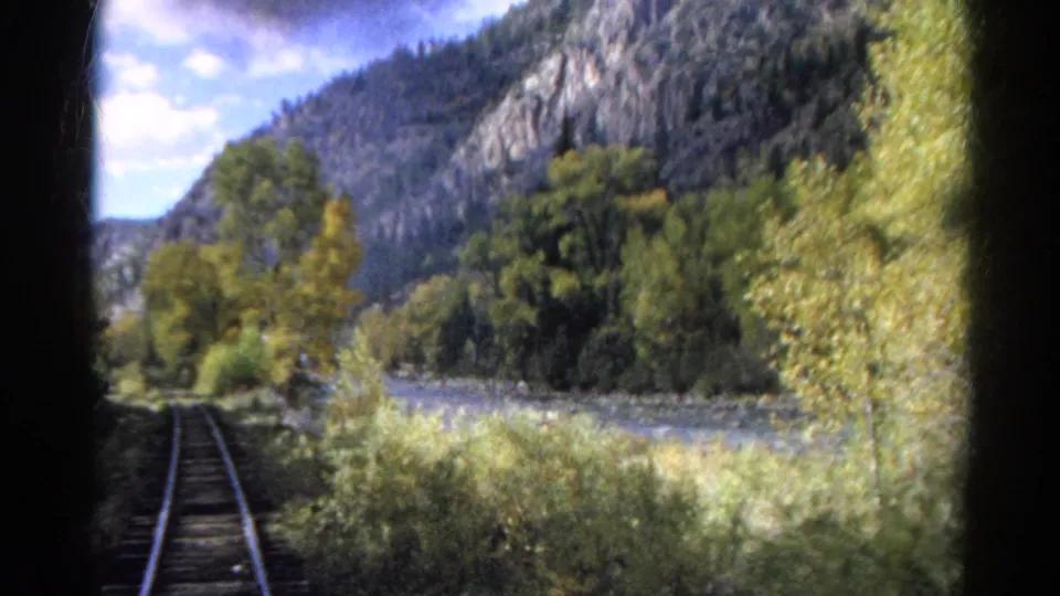 1967 Colorado 8mm frame from a moving train showing tracks, riverside trees, rocky mountains, and dark film-gate edges.