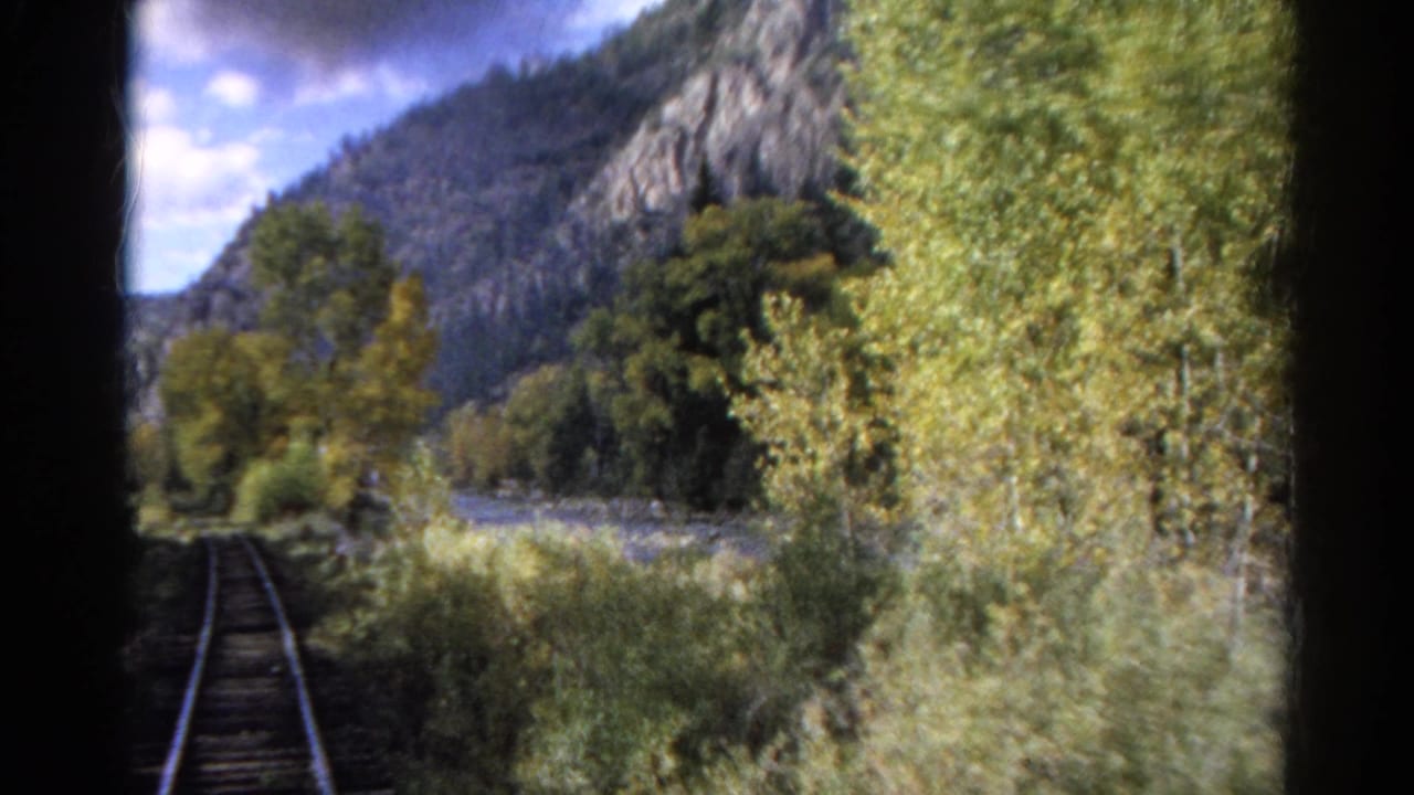 Tracks run forward beside a rocky riverbank with trees and mountain slopes ahead.