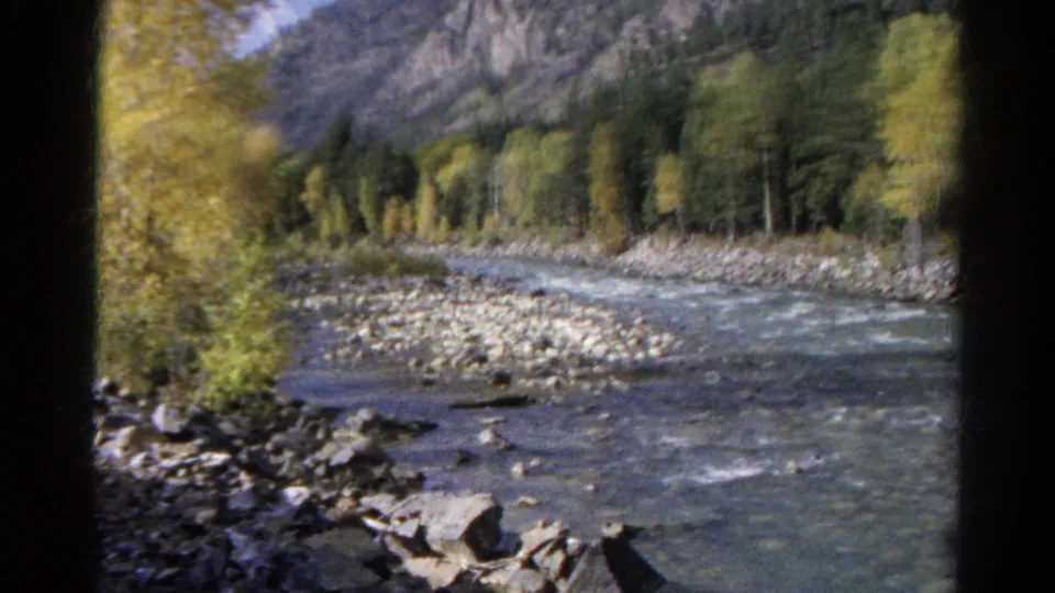 1967 Colorado 8mm frame looking from a moving train toward a rocky river, trees, and mountain backdrop.