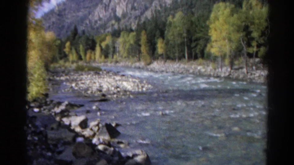 1967 Colorado home-movie frame showing a rocky river flowing below trees and mountain slopes from a train-side viewpoint.