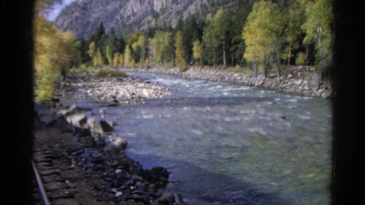 River and tree-lined bank viewed from a train-side position with mountains beyond.