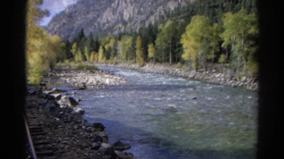 A soft 8mm view of a Colorado river bend from a moving train, with track edge, rocks, trees, and mountain terrain visible.