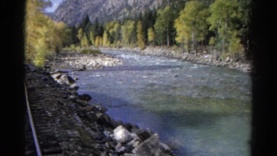 Final 1967 Colorado frame showing the river beside train tracks, with rocks in the foreground and trees lining the mountain valley.