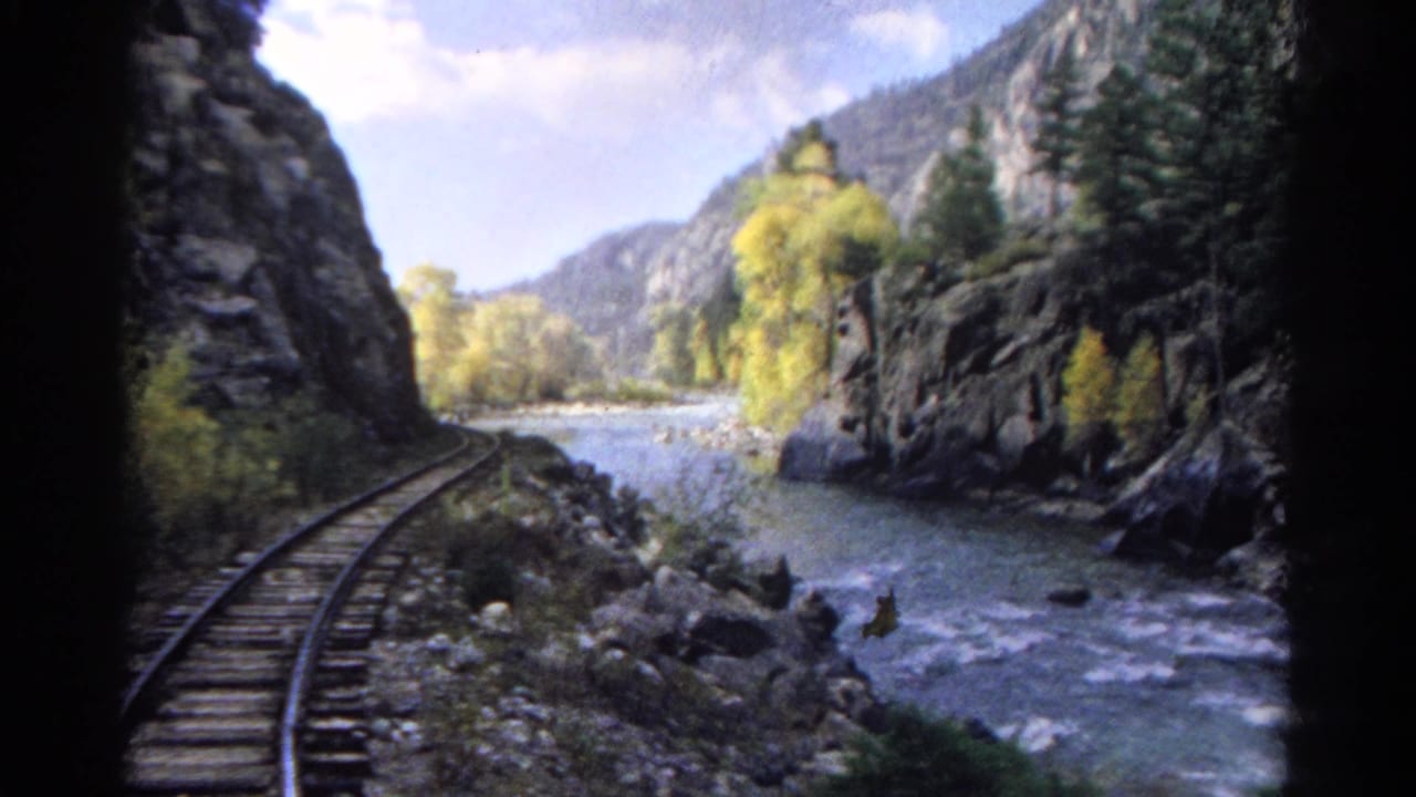 Colorado train-window view from 1967, with track, river, rocky slopes, evergreens, and yellow foliage visible ahead.