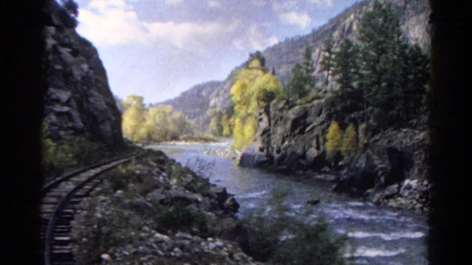Moving 8mm train view in Colorado, looking past a curved rail line toward a river, rock walls, and fall-colored trees.