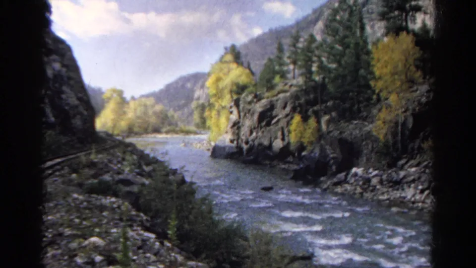 1967 Colorado home-movie frame from a train, showing a river bend, rocky canyon sides, evergreens, and yellow autumn foliage.