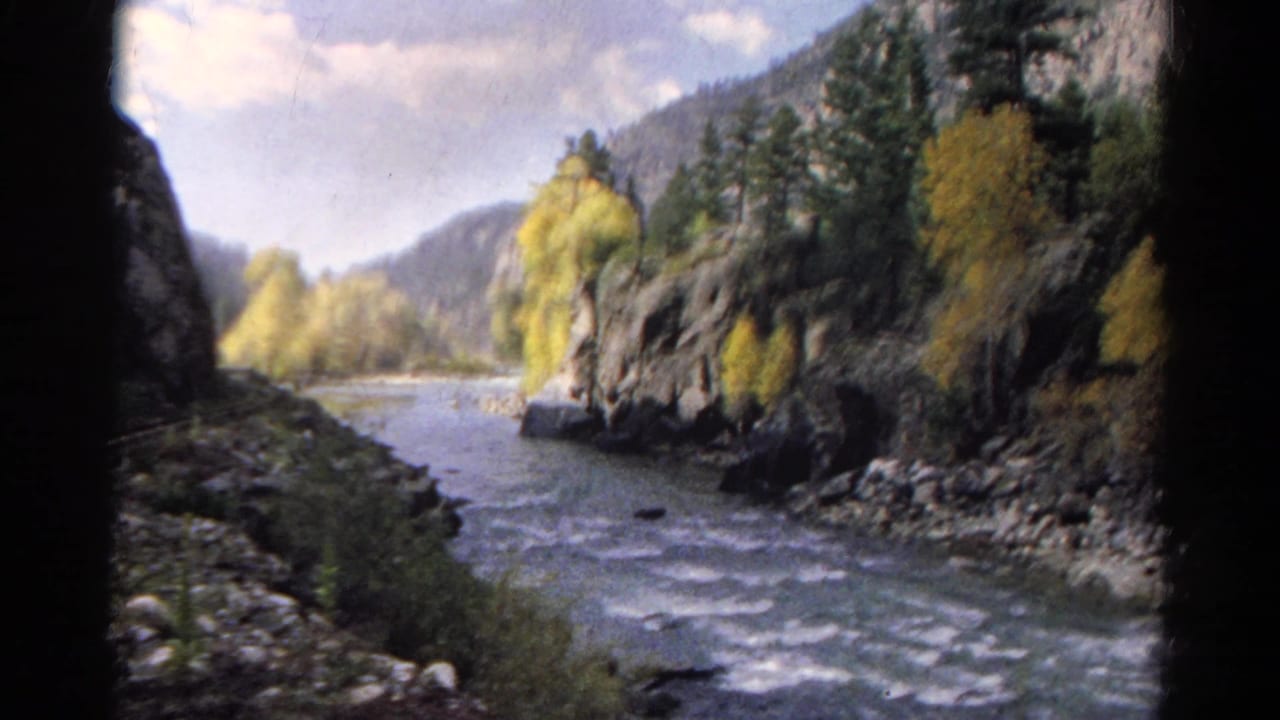 Rushing river between rocky banks and forested mountains, seen through dark train-window framing.