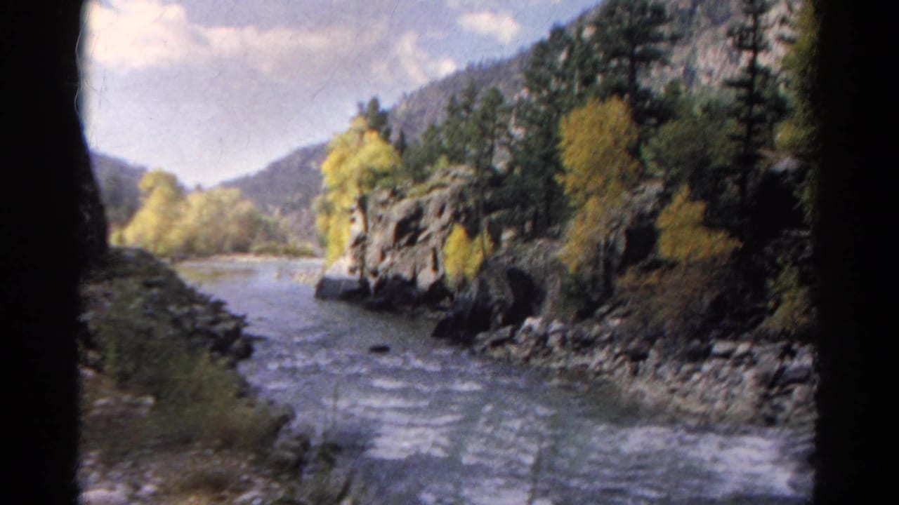 1967 Colorado home-movie frame showing a river beside rocky slopes, evergreens, and autumn yellow foliage from a moving train perspective.