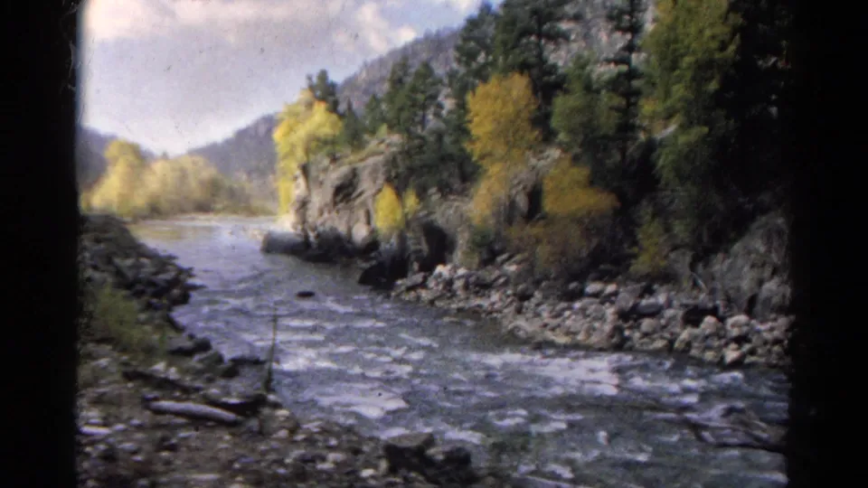 1967 Colorado 8mm travel view of a broad river channel, rocky banks, trees with autumn color, and surrounding mountain slopes.