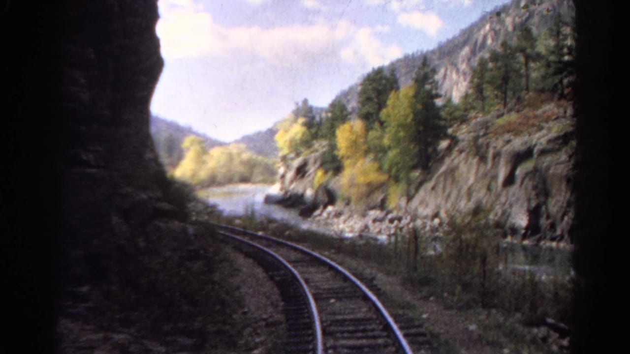 Railroad tracks curve beside a rocky river through a mountain valley.