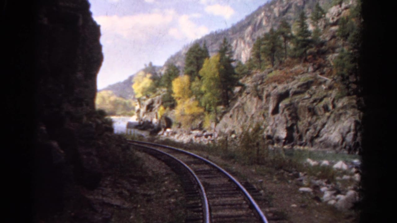 Curving train tracks run along a river below cliffs and trees.