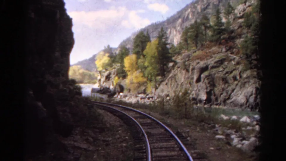Colorado mountain rail view with tracks curving beside water, cliffs, evergreens, and autumn-colored trees in 1967 8mm footage.