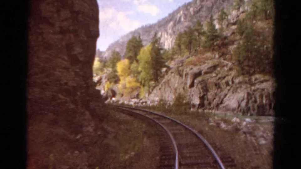 Passenger-view 8mm frame of Colorado rail tracks bending past a cliff face toward river water and mountain trees.