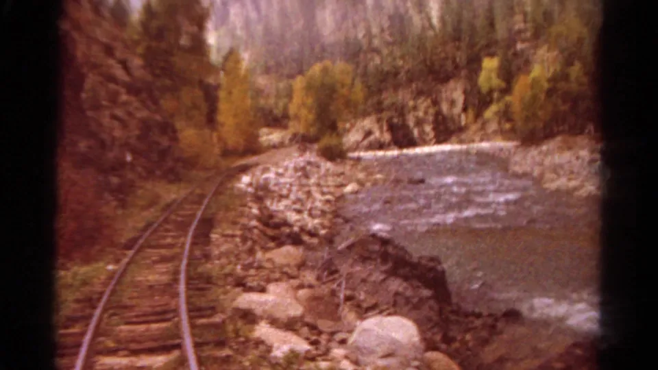 Warm orange-shifted 1967 Colorado 8mm frame showing railway tracks beside a rocky riverbank and canyon trees.
