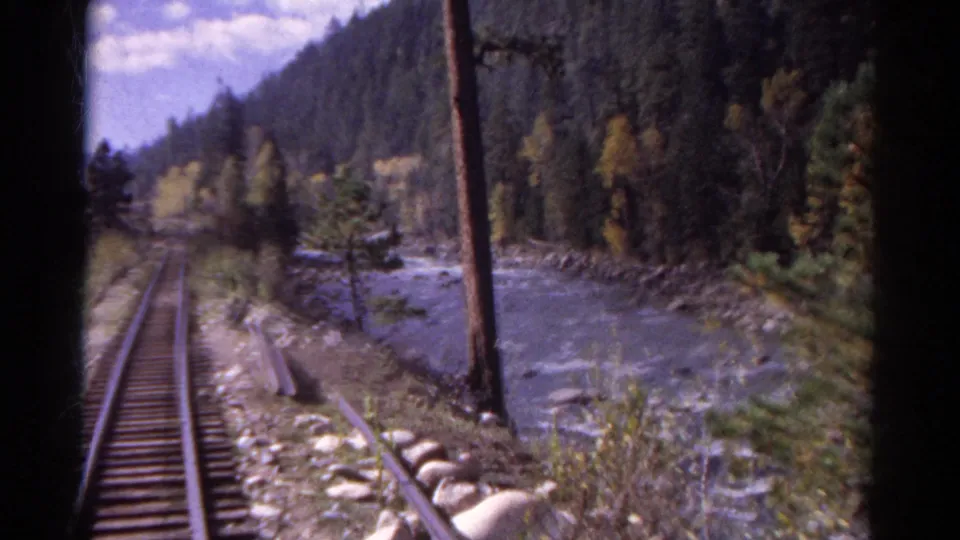 1967 Colorado train-track view beside a mountain river, with rails, utility pole, trees, and rocky shoreline.