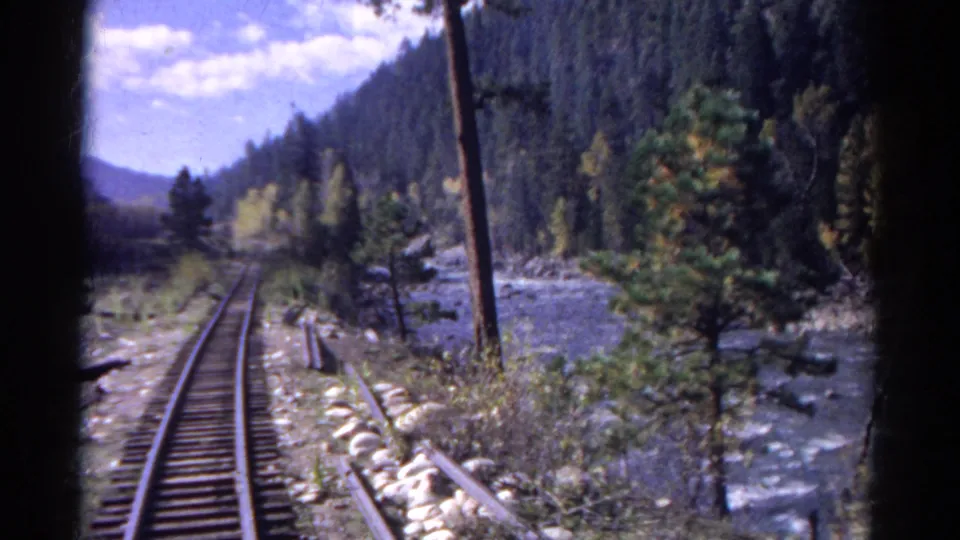 Colorado 1967 8mm frame looking along railroad tracks beside a river, with forested slopes and brush in the foreground.
