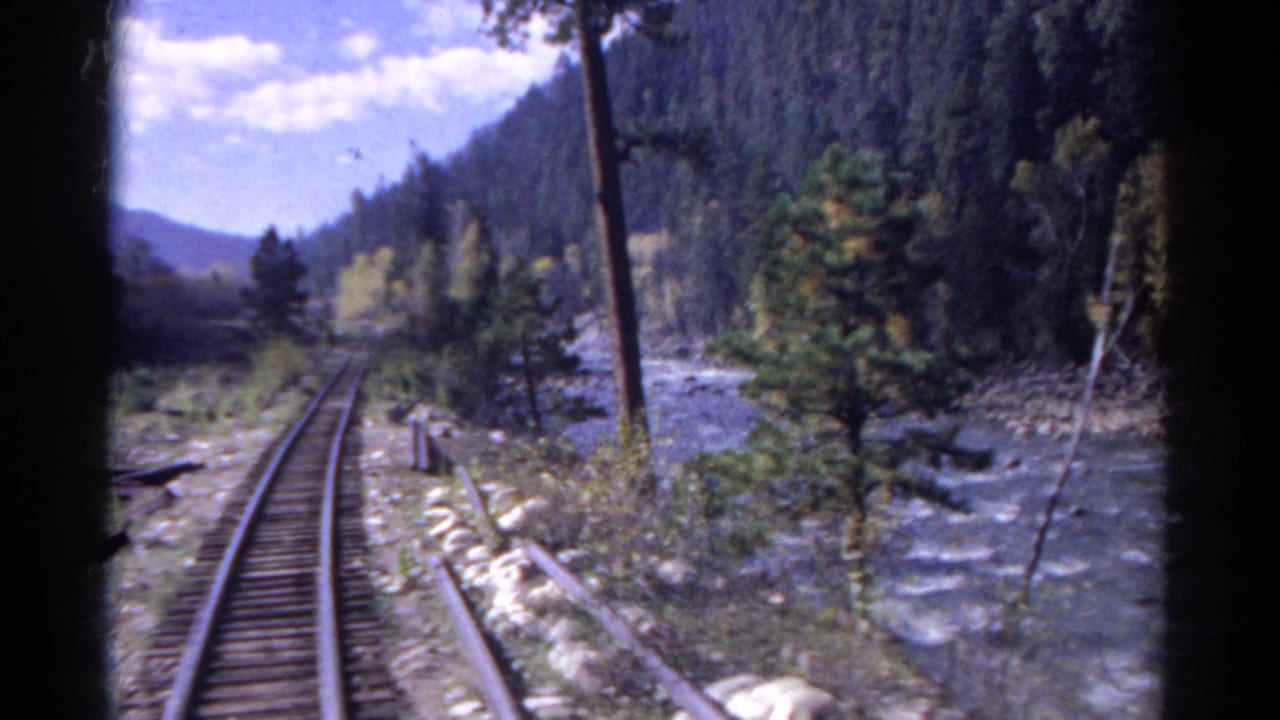 1967 Colorado 8mm frame from a moving train, showing curving tracks beside a rushing river and wooded hills.