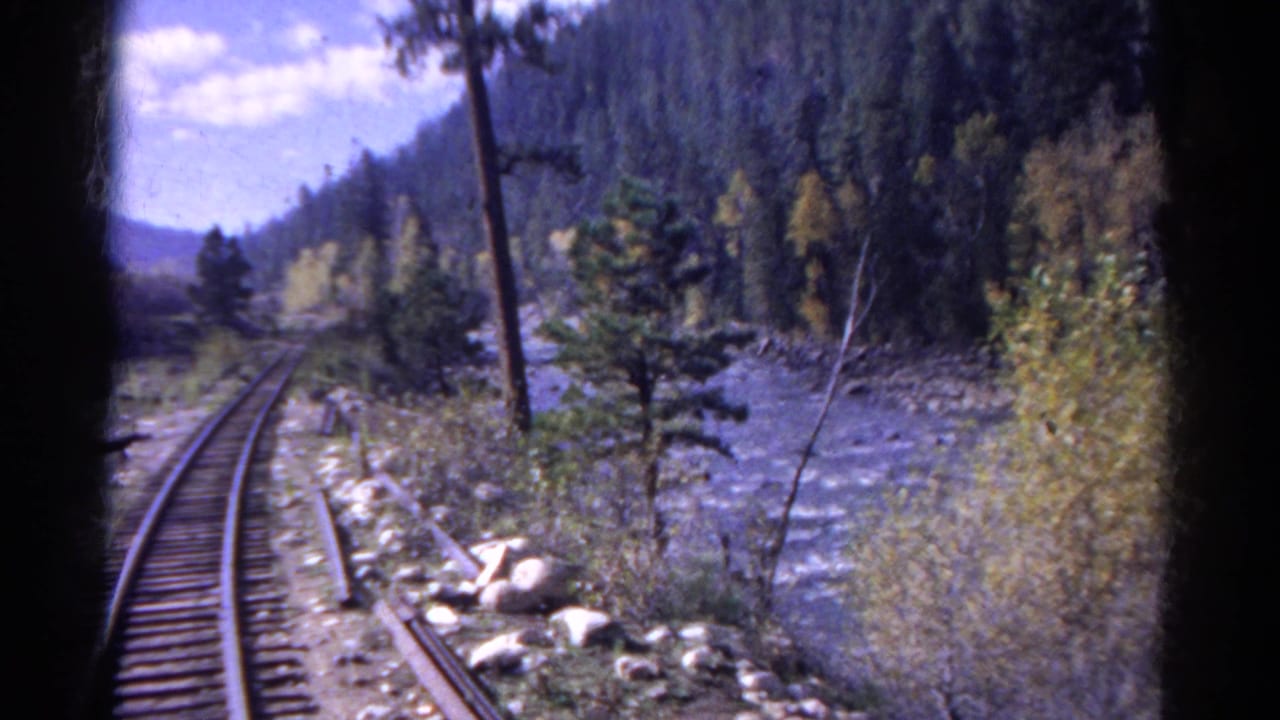 Passenger-view frame of Colorado rail tracks following a rocky river through evergreen hills and yellowing trees.