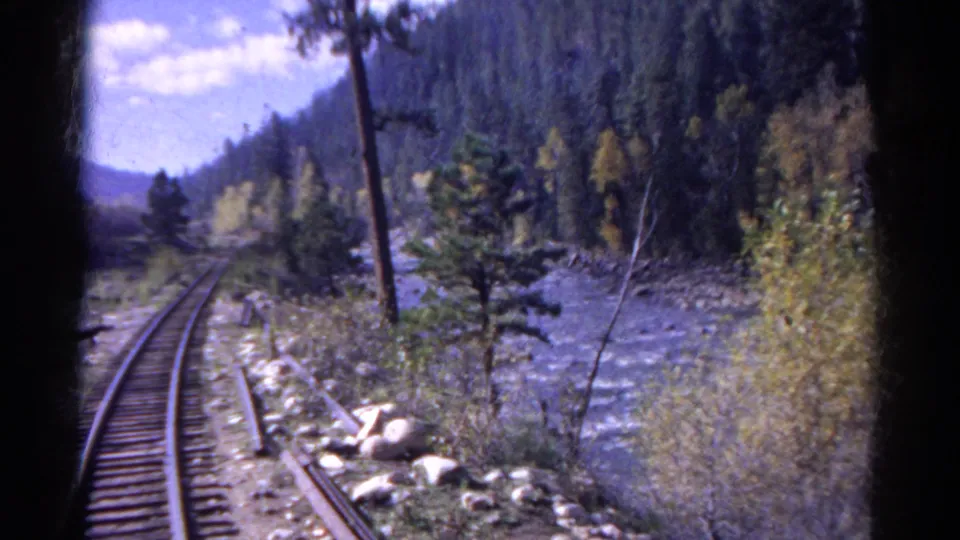Passenger-view frame of Colorado rail tracks following a rocky river through evergreen hills and yellowing trees.