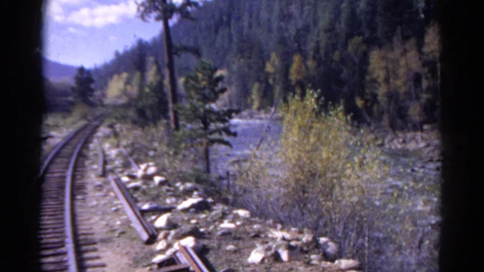 1967 home-movie frame looking forward from a train as rails curve past brush, rocks, and a mountain river in Colorado.