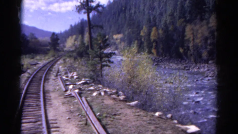 End frame of a 1967 Colorado train view, with the track curving beside a rocky river and dense hillside trees.